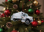 A white garbage truck Christmas ornament decorated with colorful holiday string lights, hanging by a red ribbon on a white background.